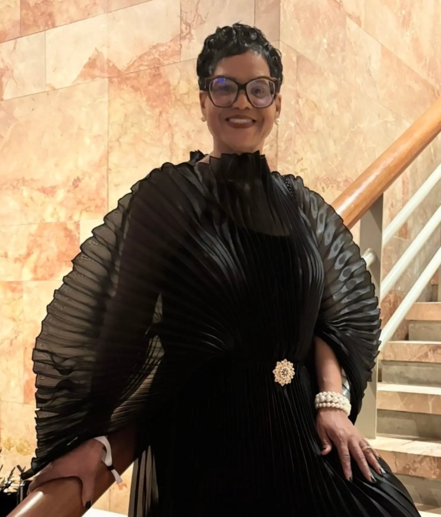 Dr. Lucretia Mason-Underdue in a black pleated gown on a marble staircase